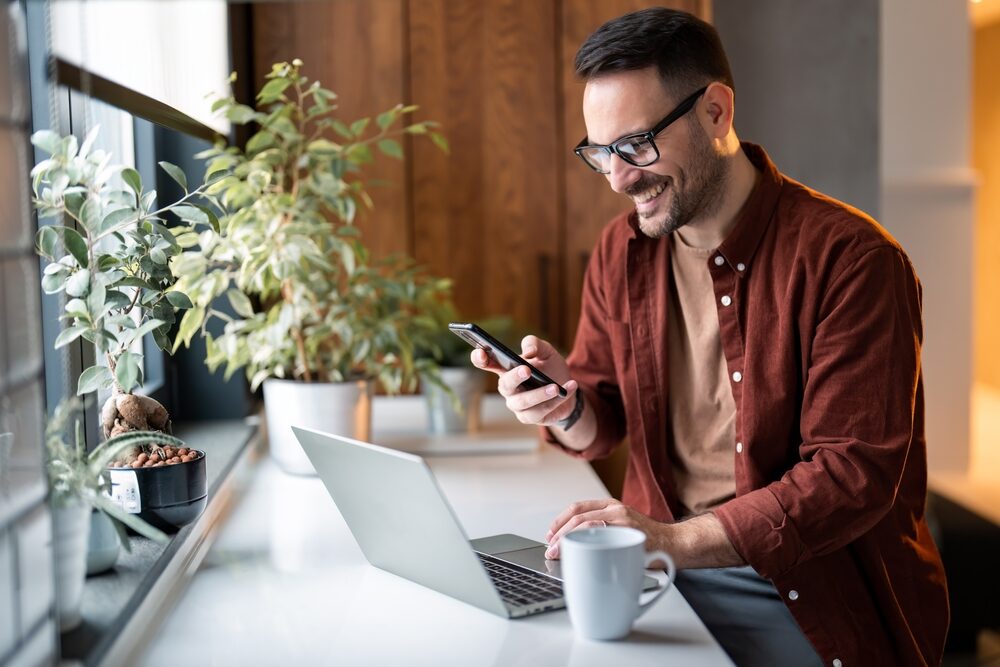 Smiling man using laptop and smartphone near a sunny window, representing convenience in selling homes online – Sell My House Oceanside
