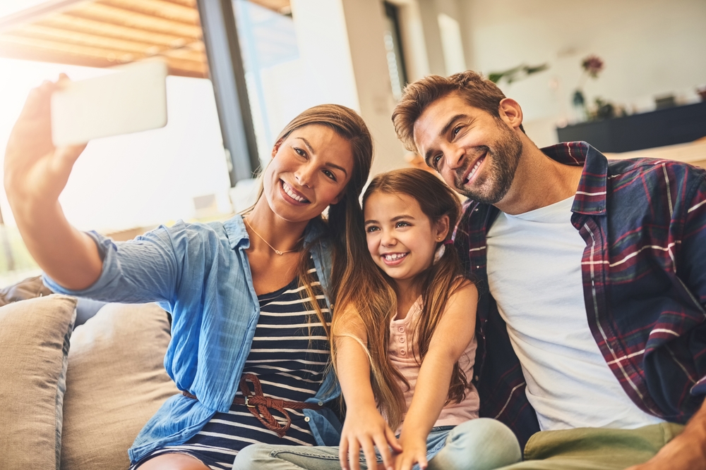 Happy family sitting together on the couch taking a cheerful selfie after selling their home – Sell My House Oceanside