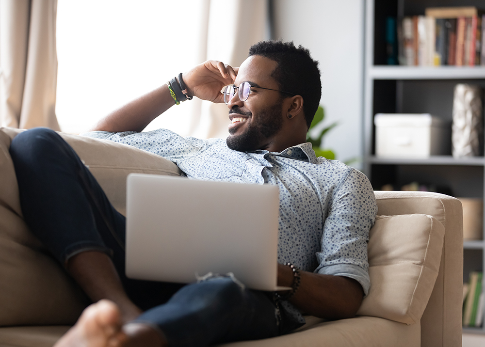 Man relaxing on couch with laptop – King Beach Residential Smiling man in glasses reclines comfortably on a beige sofa with a laptop on his lap, enjoying a relaxed moment at home with bookshelves in the background – King Beach Residential